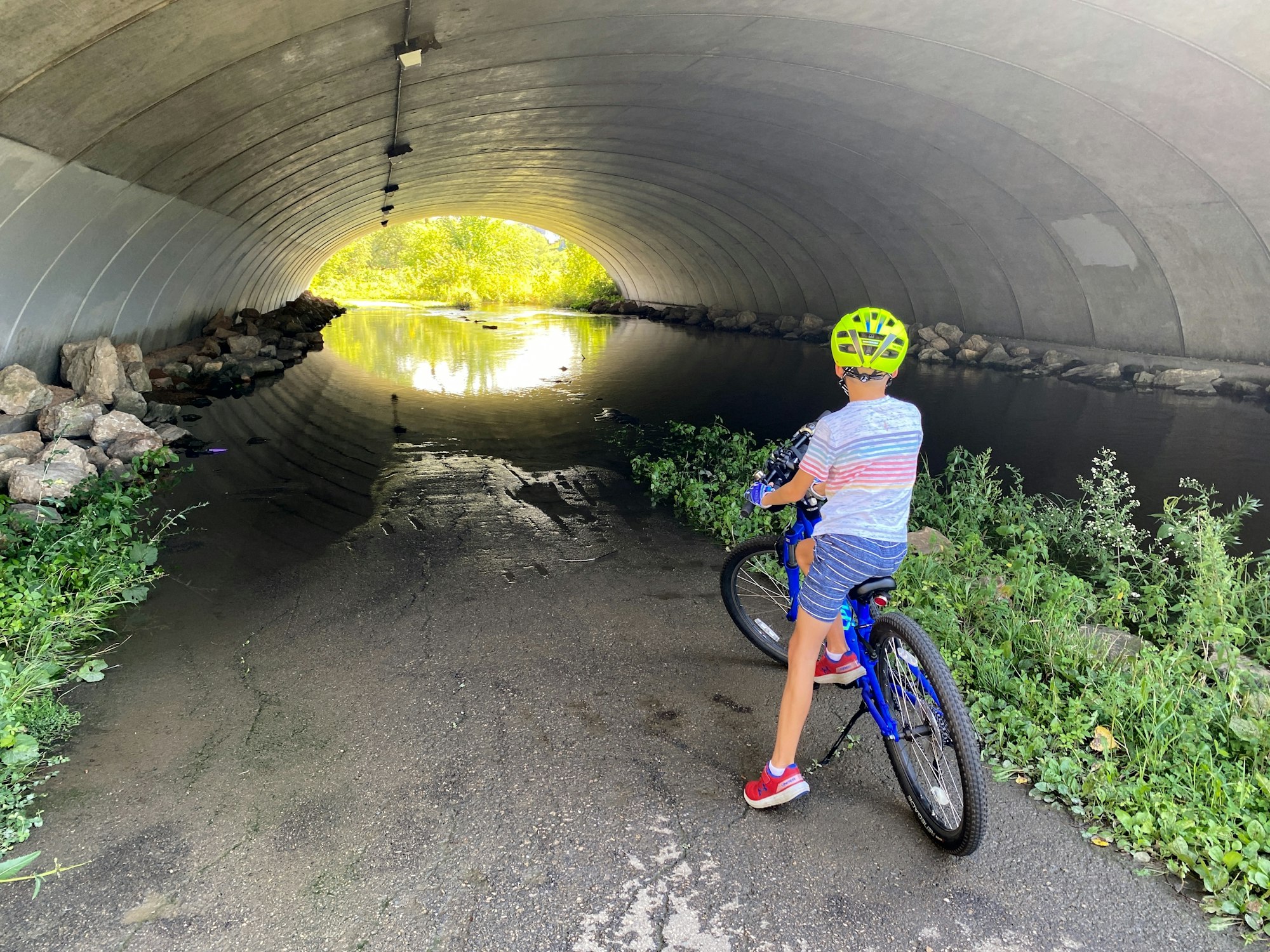 A person with a bicycle stands inside a tunnel, facing a waterway leading out to a bright, tree-lined area.