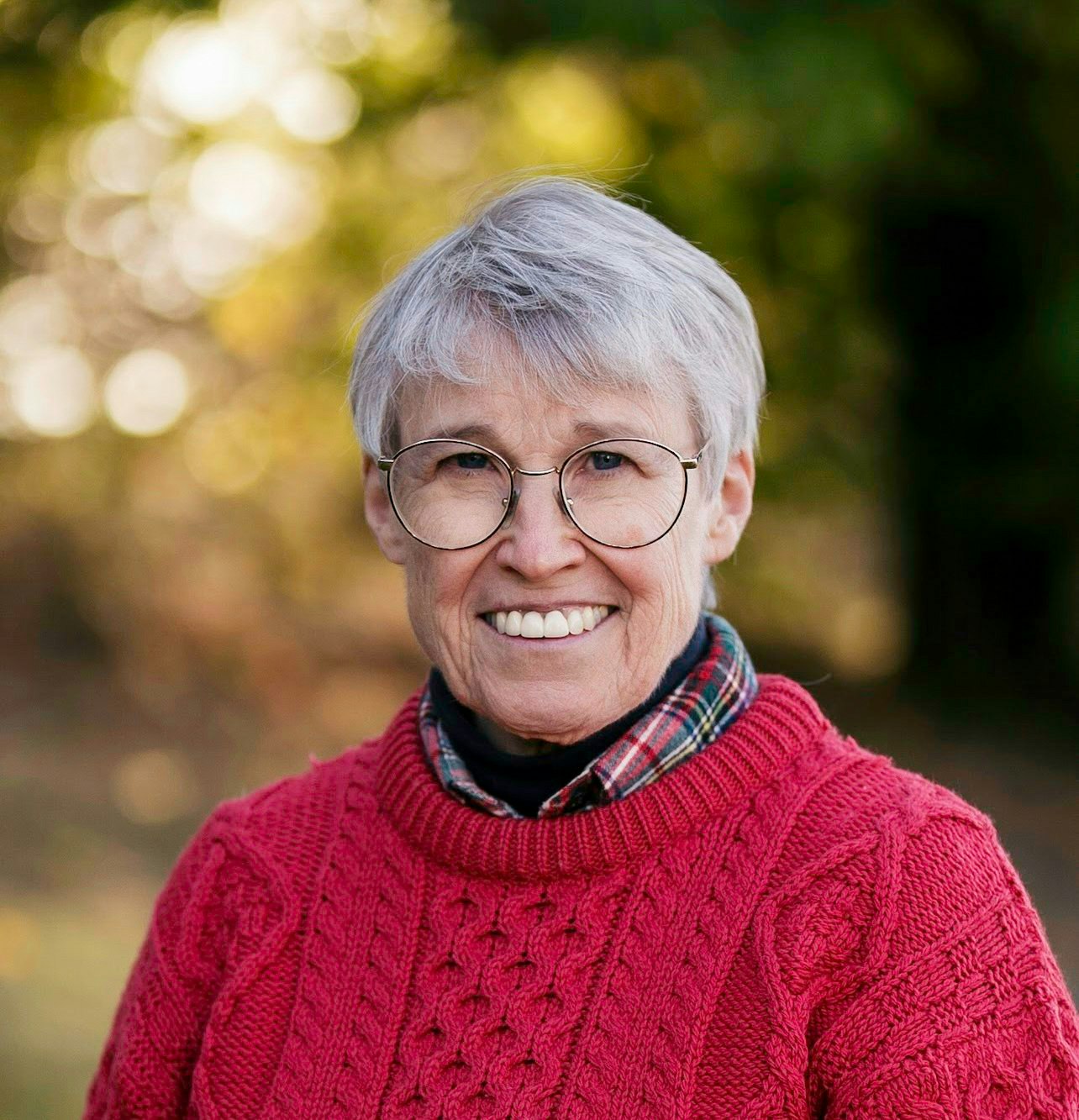 An older person wearing glasses and a red sweater, smiling outdoors.