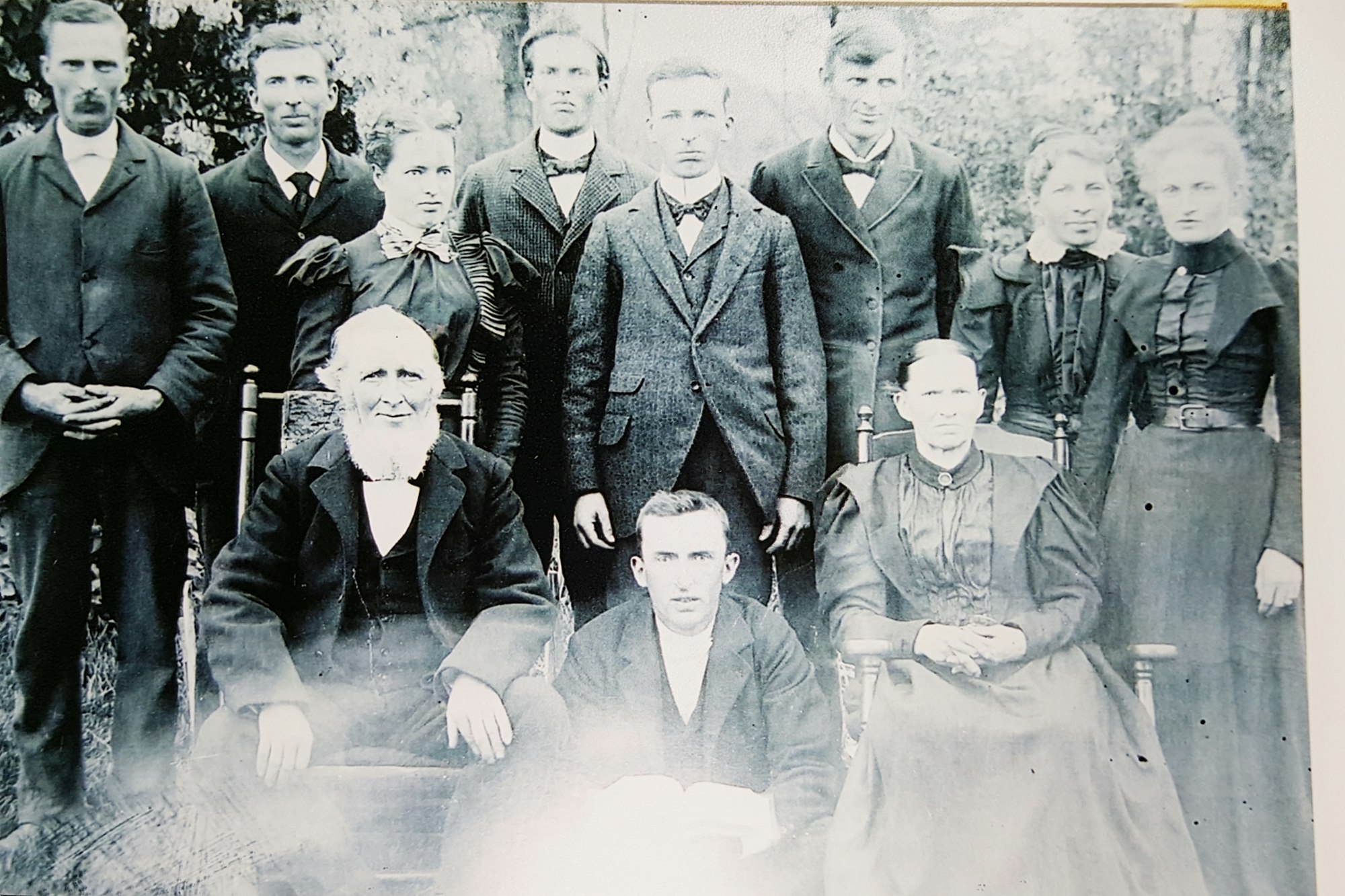 Old family photo with 9 people, seated elderly couple in front, sepia tones, vintage clothing.
