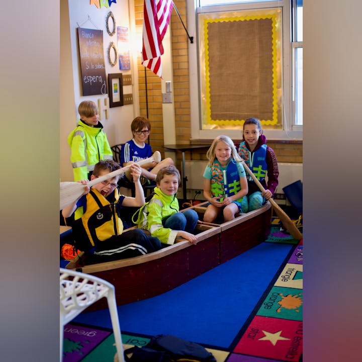 Children in life jackets sitting in a canoe indoors, smiling and holding paddles.