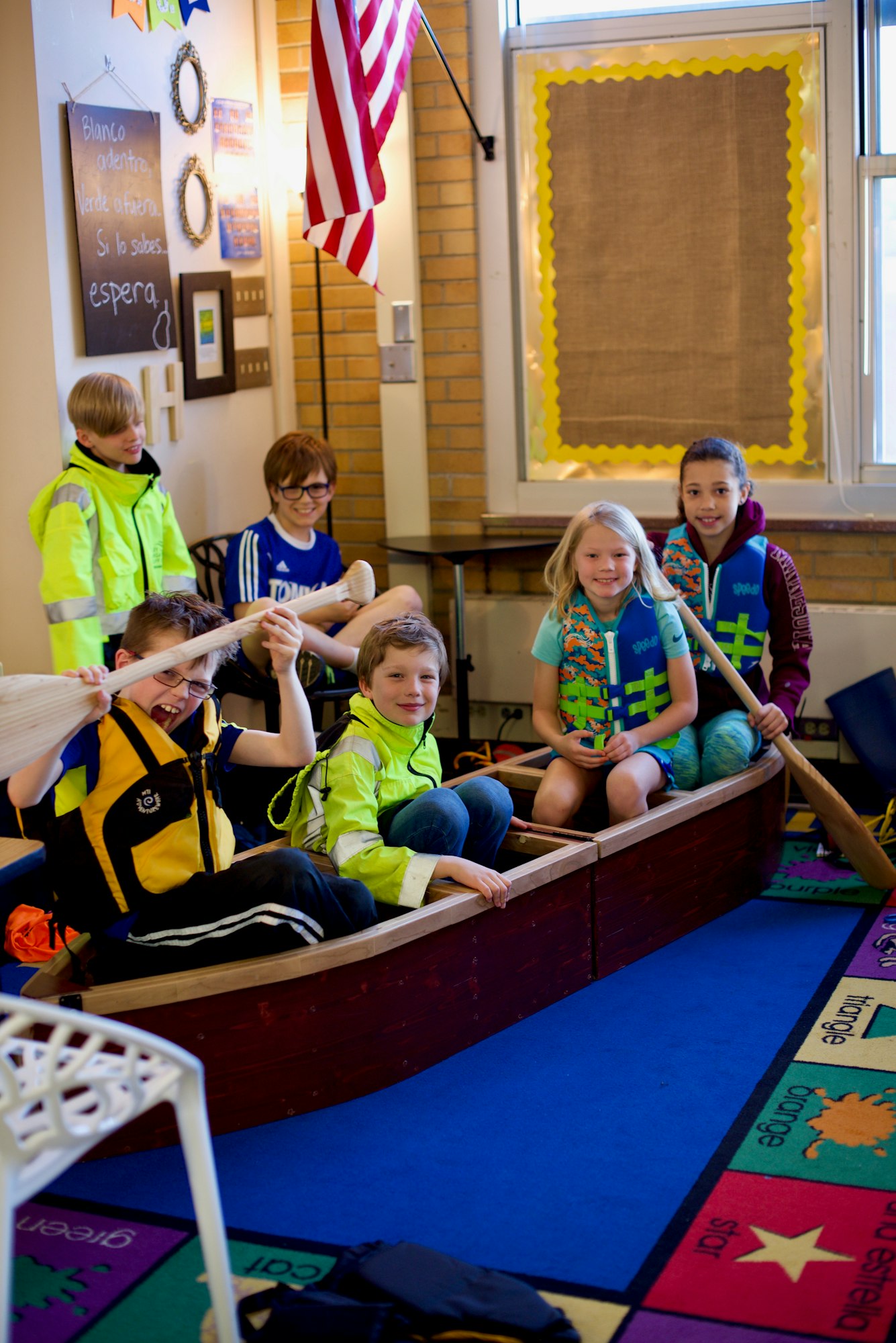 Children in life jackets sitting in a canoe indoors, smiling and holding paddles.