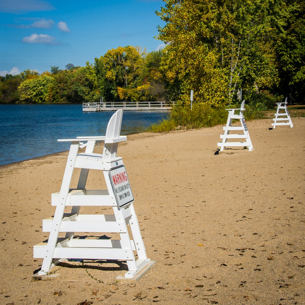 A lakeside with white lifeguard chairs, "No Lifeguard on Duty" sign, sandy beach, trees, and a pier on a sunny day.