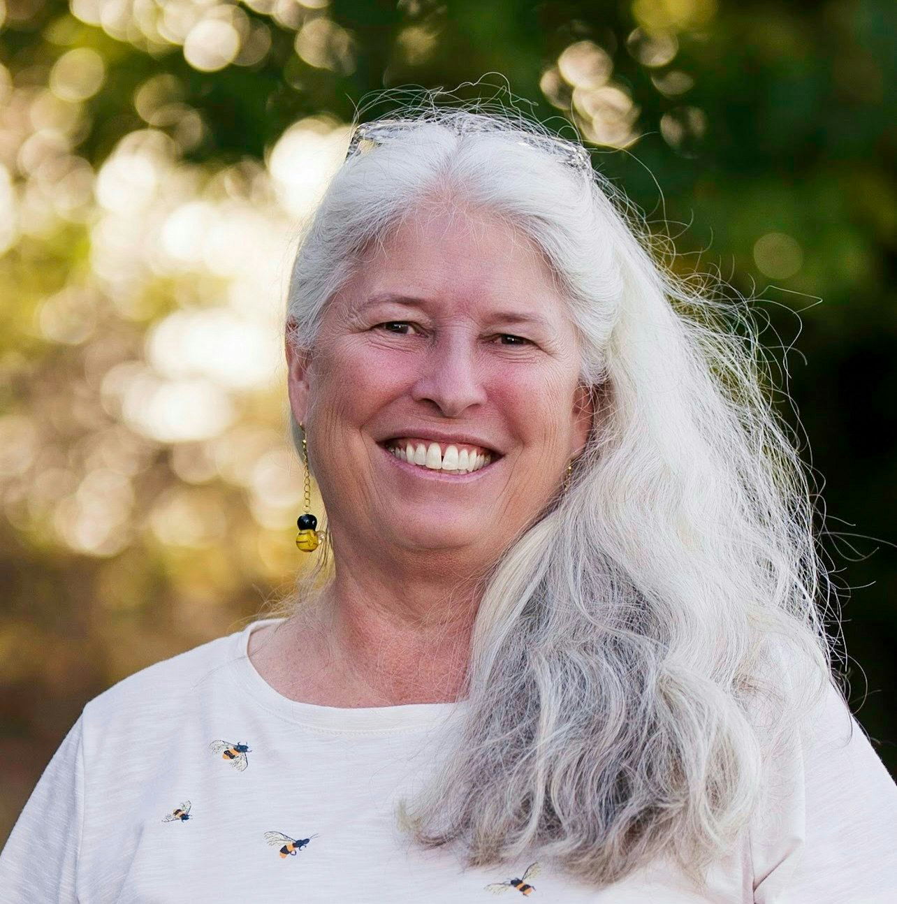 A person with long gray hair smiles outdoors, wearing a white shirt with small designs and bee-themed earrings.