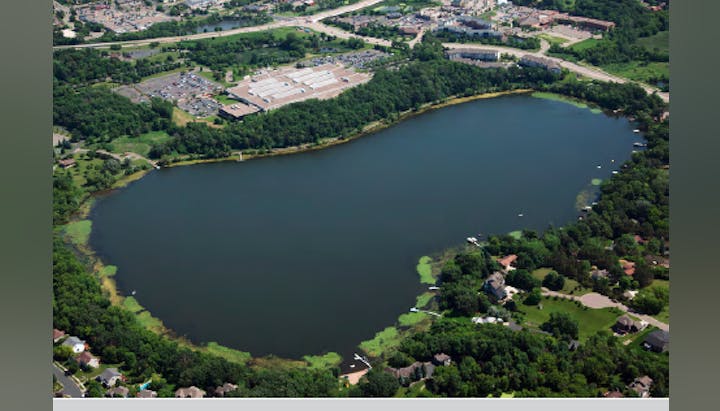 Aerial view of a lake with surrounding greenery, residential areas, and nearby industrial buildings.