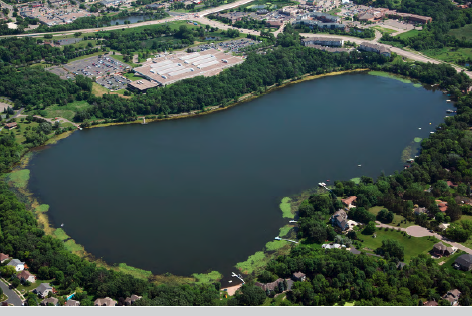 Aerial view of a lake with surrounding greenery, residential areas, and nearby industrial buildings.