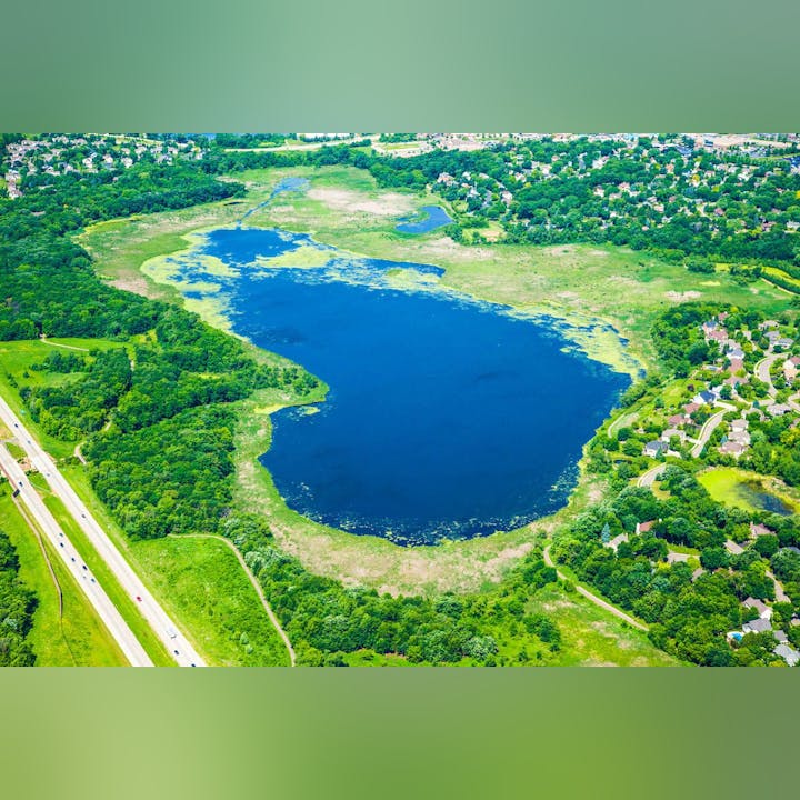Aerial view of a vibrant blue lake surrounded by greenery and residential area next to a highway.
