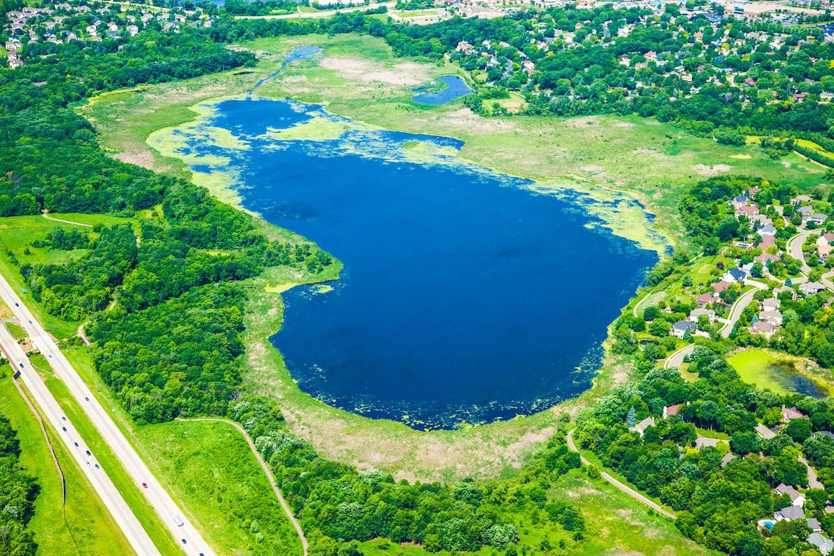 Aerial view of a vibrant blue lake surrounded by greenery and residential area next to a highway.