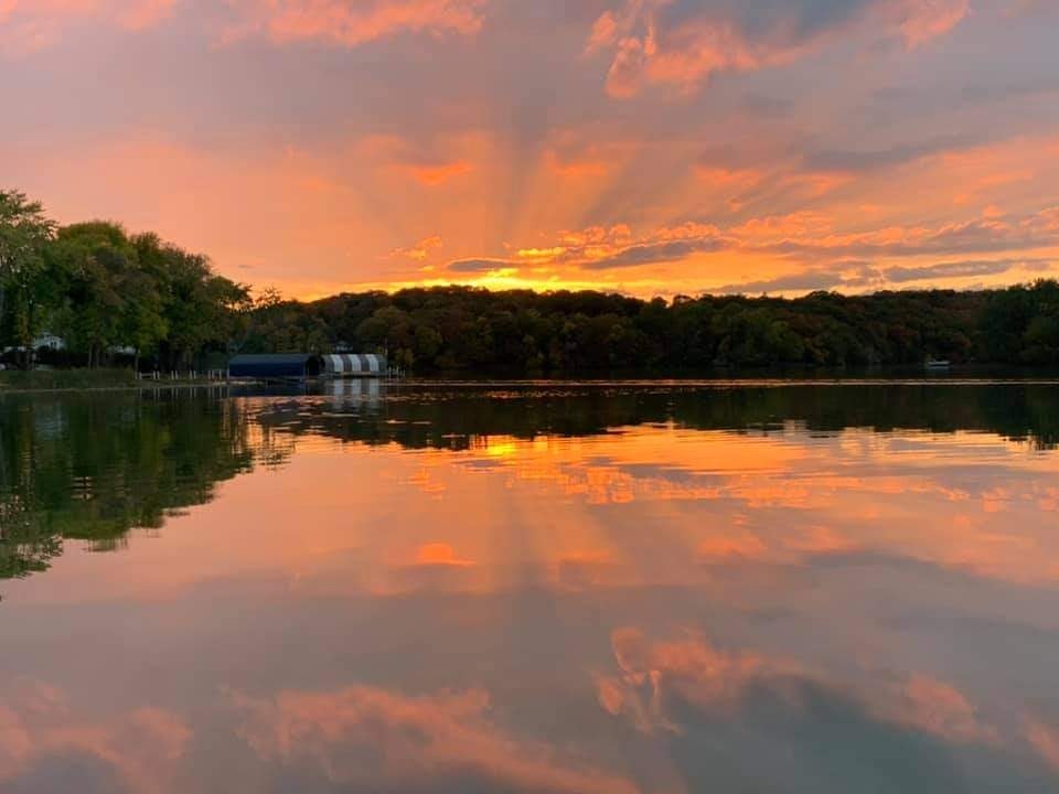 Sunset over a calm lake with colorful skies and trees reflected on the water's surface.