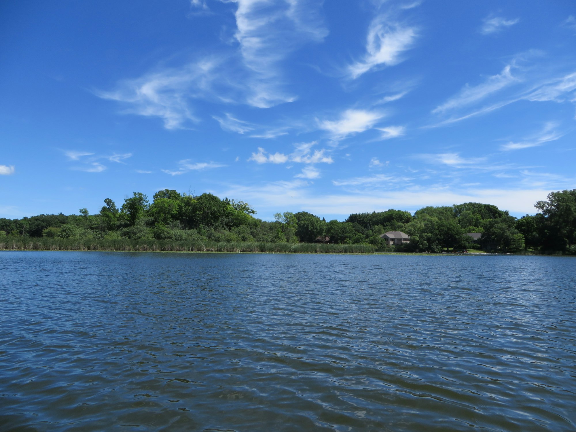 Blue sky with wispy clouds over a tranquil lake with lush greenery and a house in the distance.