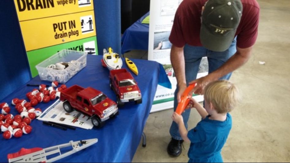 An adult and a child at an informational booth with toys and safety helmets on a table.