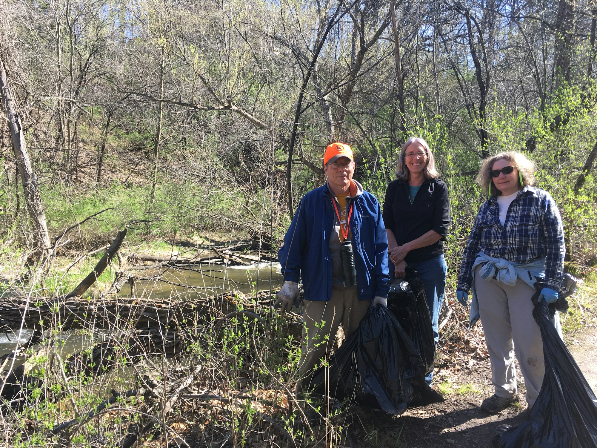 Three people with trash bags in a wooded area, possibly after a clean-up activity.