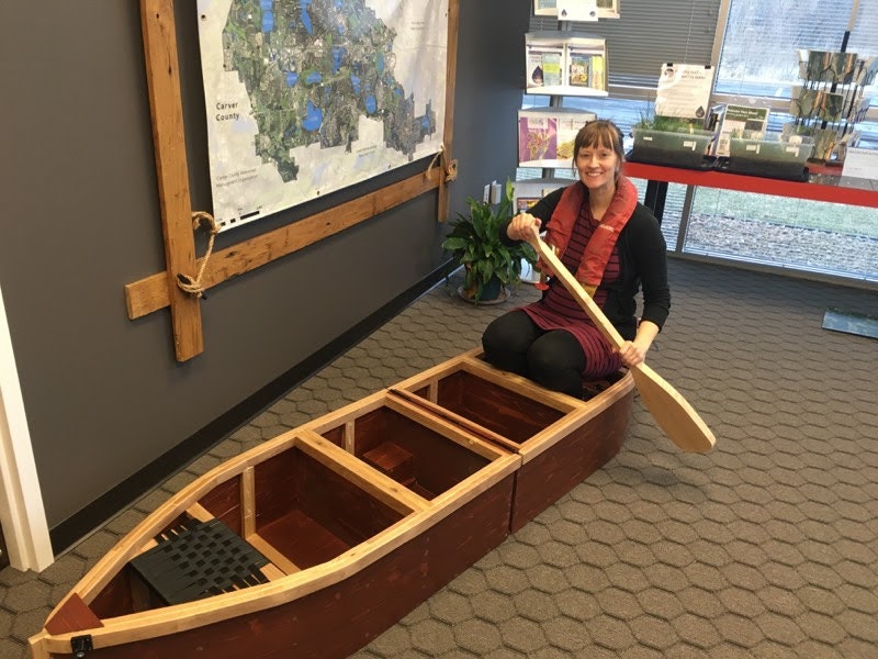 A smiling person sitting in a canoe indoors, holding a paddle, with a map in the background.