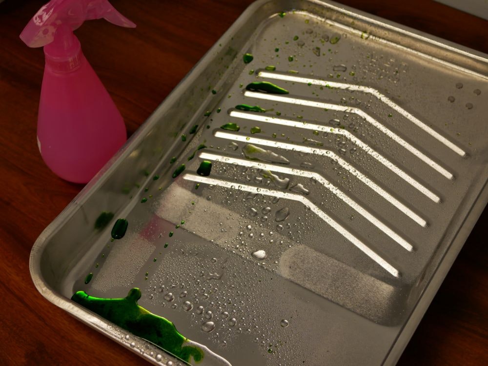 A pink spray bottle and green liquid droplets on a silver metal tray with water beads.