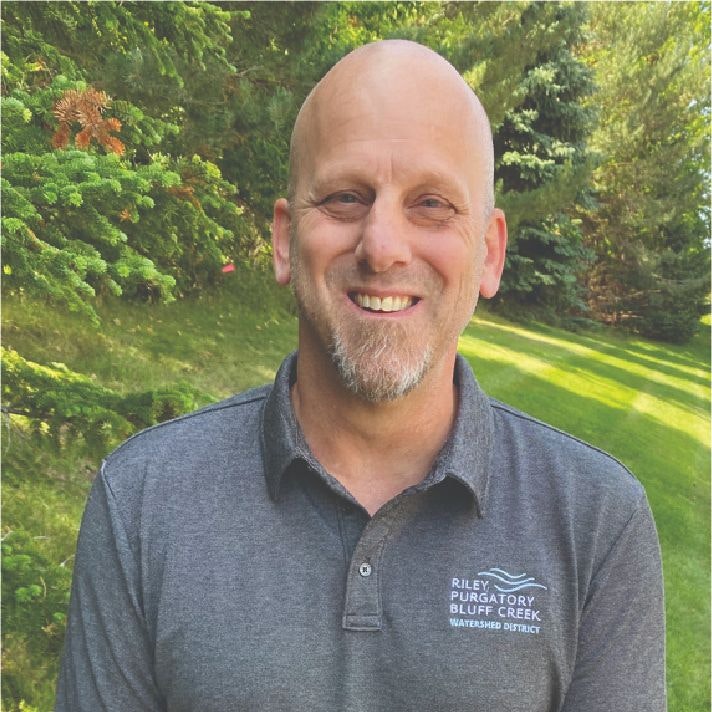 A smiling bald man in a polo shirt with a logo, outdoors with greenery in the background.