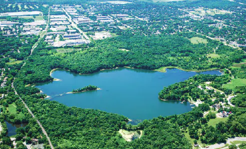 Aerial view of a lake surrounded by greenery with urban areas nearby.