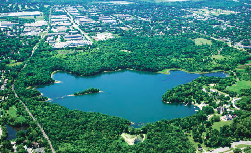 Aerial view of a lake surrounded by greenery with urban areas nearby.