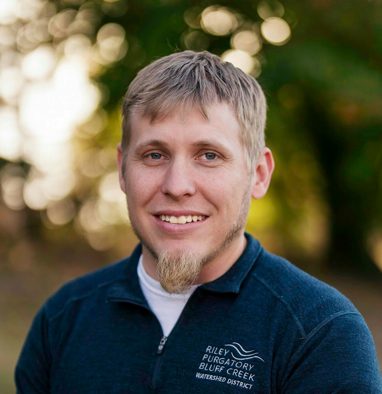 A man with short hair and a goatee, wearing a dark shirt with text, smiling outdoors with blurred trees in the background.