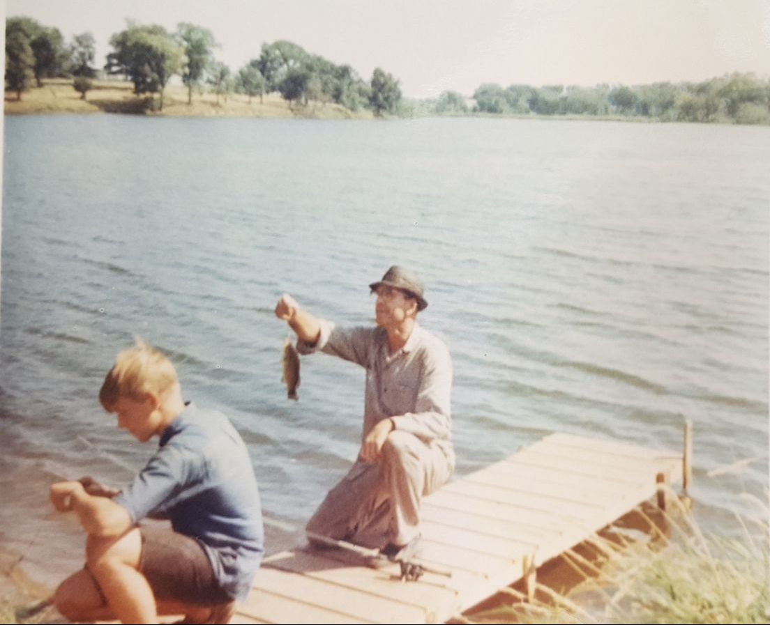 Two people on a wooden dock by a lake, one holding a fish. Vintage photo.