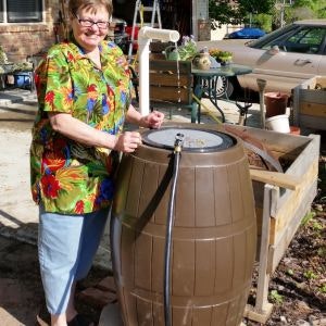A smiling person beside a brown rain barrel with a tap, outdoors near a driveway.