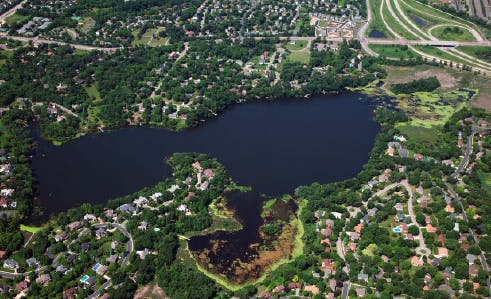 Aerial view of a lake with surrounding residential area and greenery, roads visible on the right side.