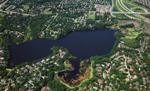 Aerial view of a lake with surrounding residential area and greenery, roads visible on the right side.