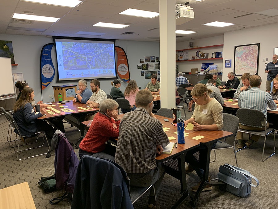 A group of people in a meeting room with a projected map, sitting at tables with notes and discussions underway.