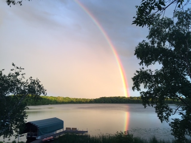 A vibrant rainbow above a serene lake with a docked boat, surrounded by lush trees.