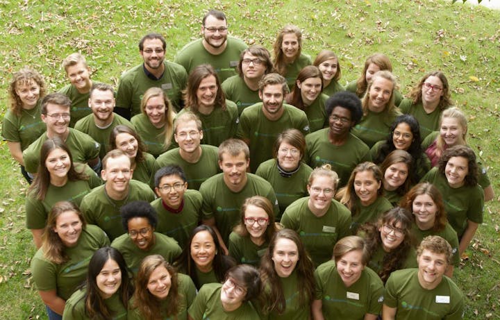 Group of smiling people in green shirts, viewed from above, standing on grass.