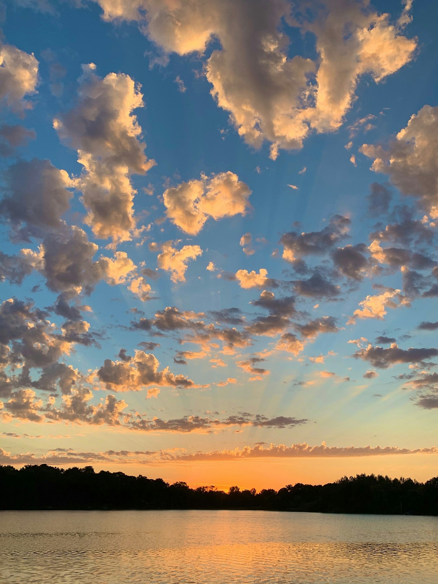 Sunset with crepuscular rays over a tranquil lake and scattered clouds in a colorful sky.
