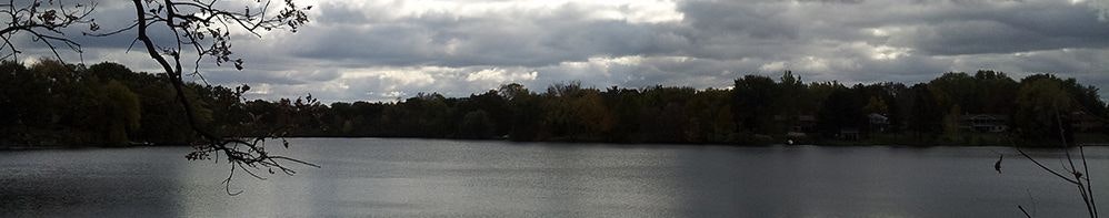 A tranquil lake under a cloudy sky with autumn trees and houses along the shore.