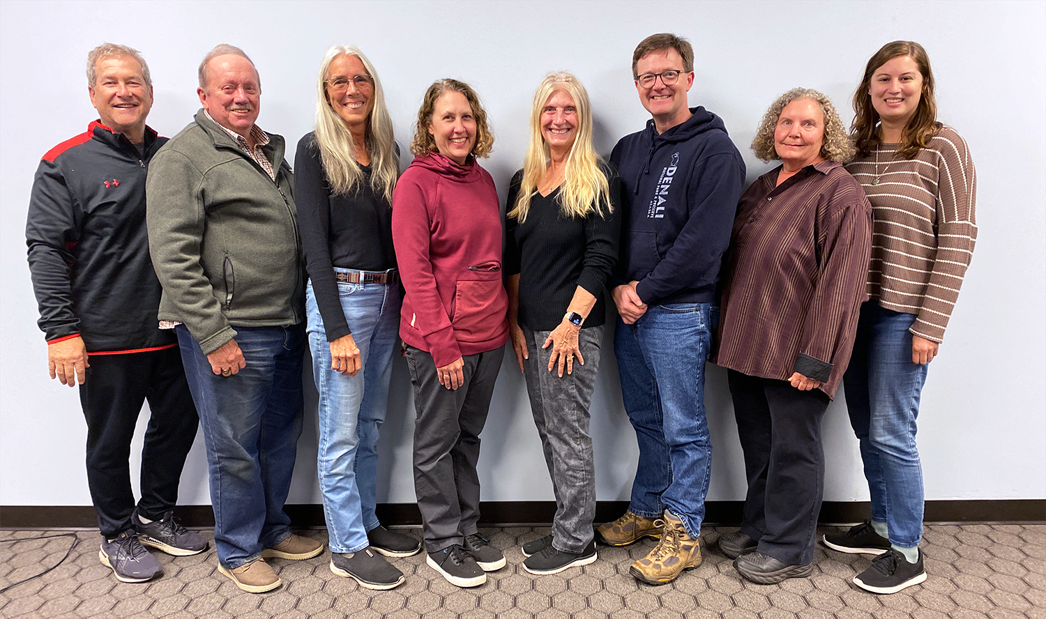 A group of eight people standing side by side in casual clothing against a plain background.