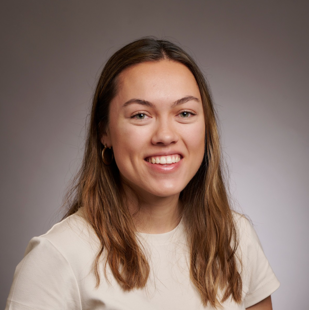 A smiling person with long, wavy hair wearing a light-colored shirt, photographed against a neutral background.