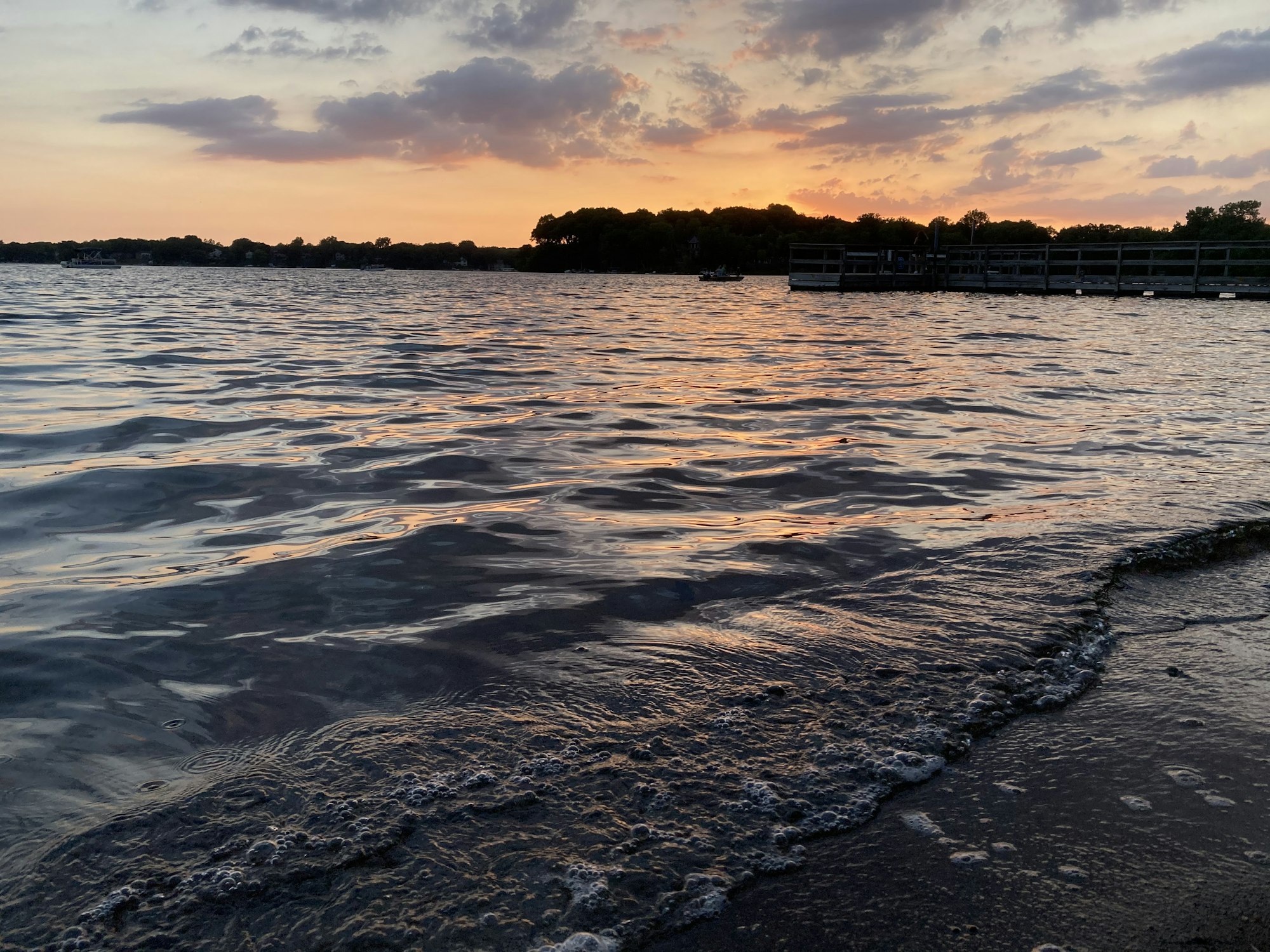 Sunset over a lake with small waves hitting a sandy shore and a dock in the background.