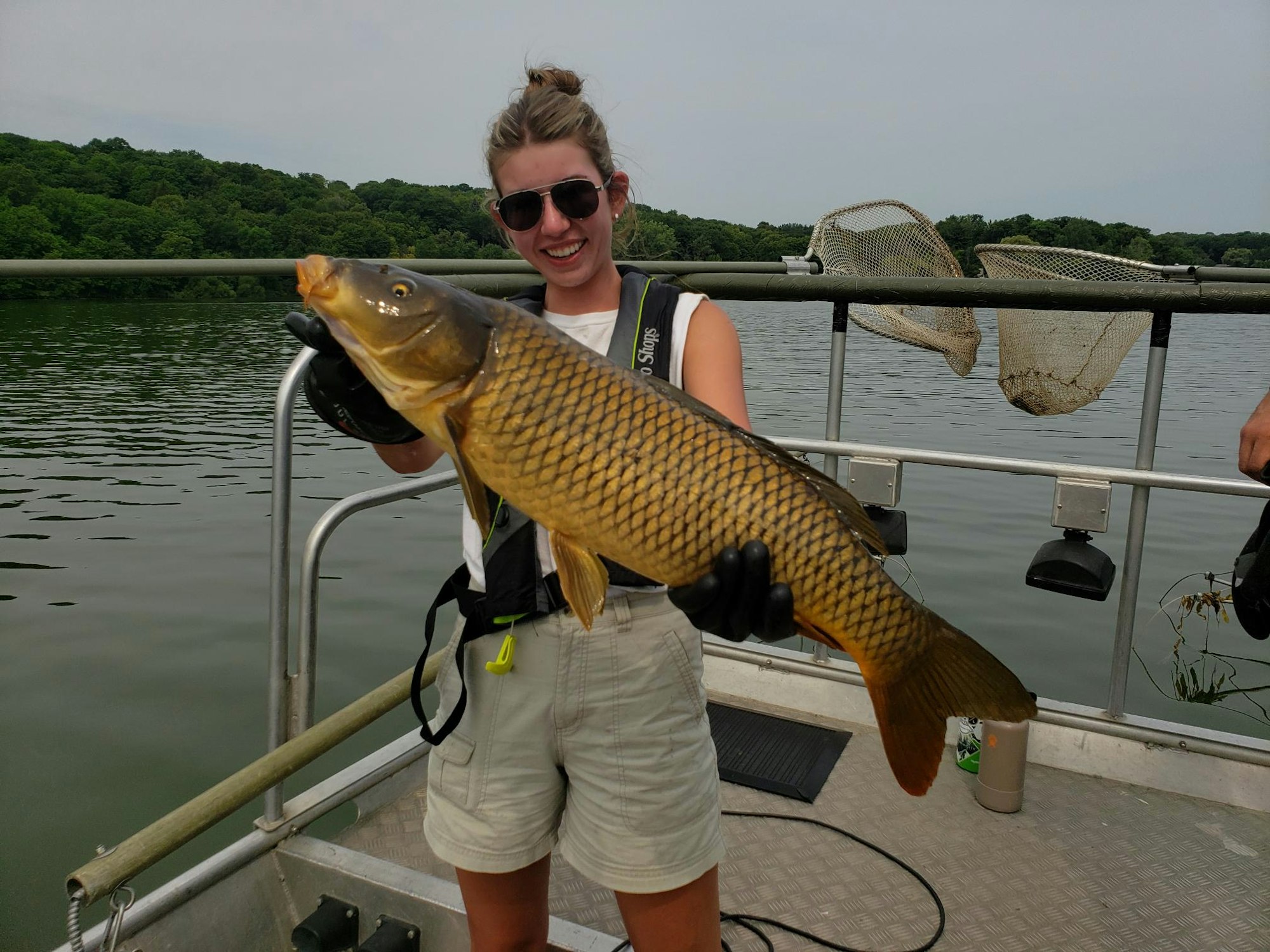 A person holding a large fish on a boat, smiling, with a lake and trees in the background.