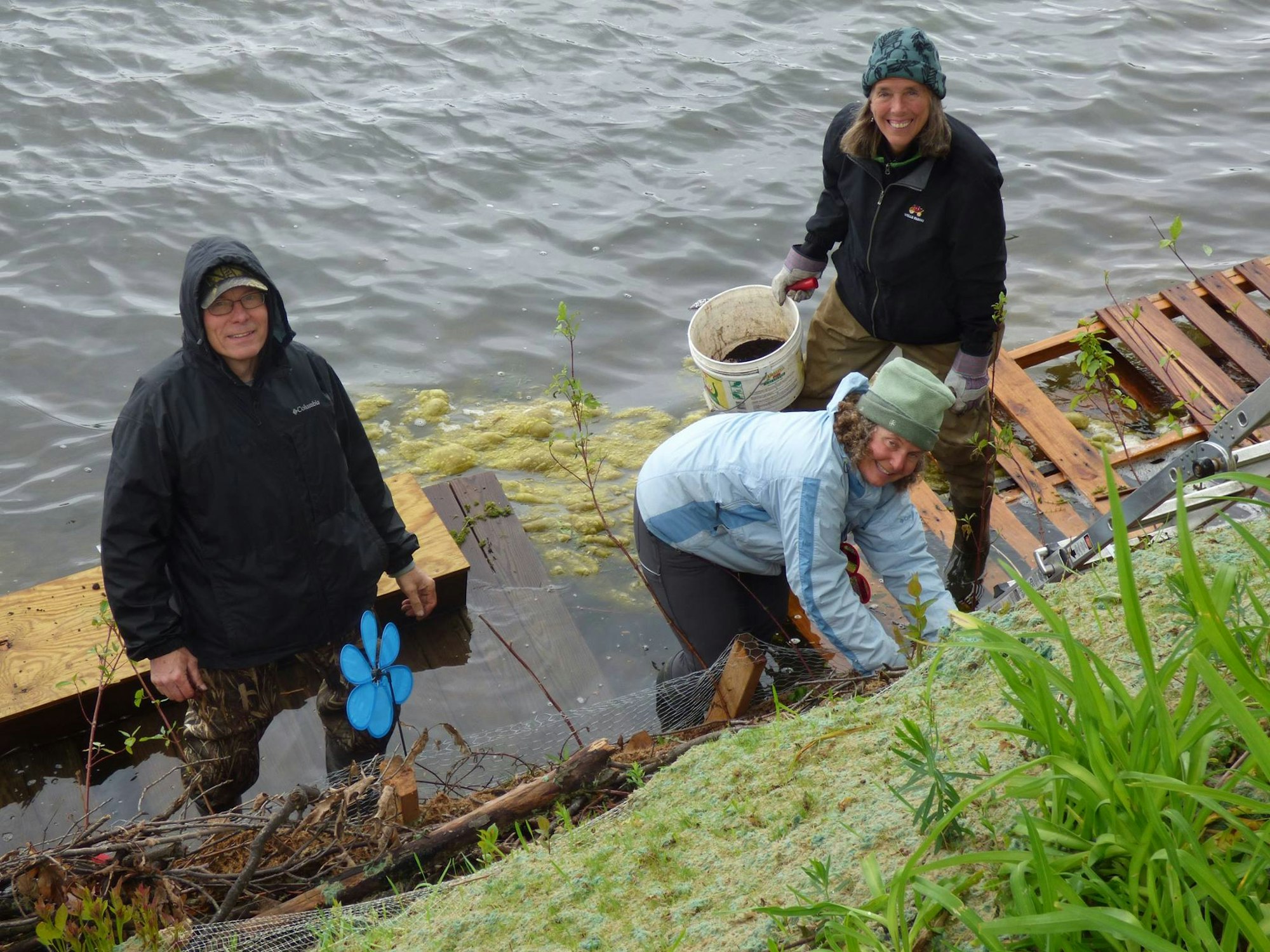Three people by a lake, working on a wooden structure, surrounded by vegetation and overcast skies.
