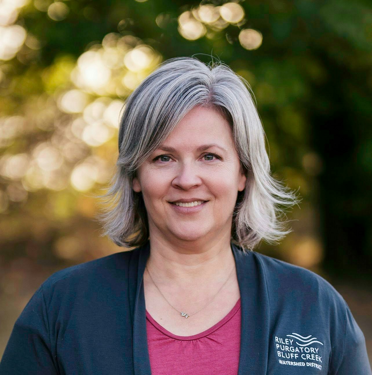 A person with gray hair smiling, wearing a dark jacket with "Riley Purgatory Bluff Creek Watershed District" written on it, outdoors.