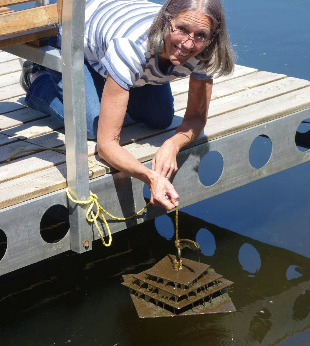 Woman kneeling on a dock, smiling and lifting a square metal structure from water using a rope.