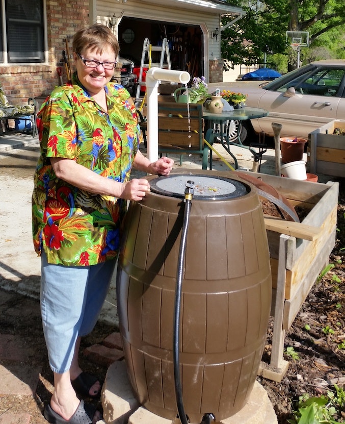 Woman with a rain barrel, colorful shirt, driveway, and part of a house.