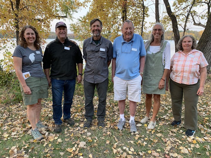 Six people standing together outdoors in autumn, with leaves on the ground and a lake in the background.