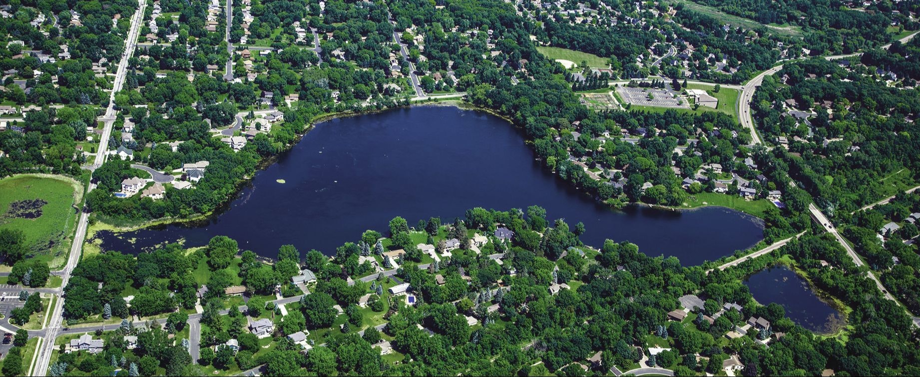 Aerial view of a suburban area with a lake, roads, houses, and greenery.