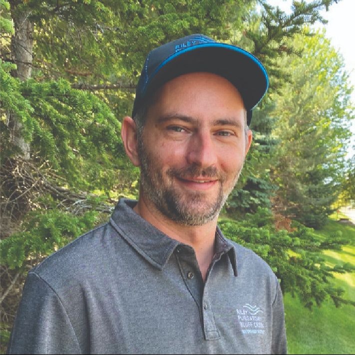 A man in a cap smiling with trees in the background.