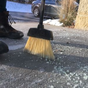 A broom, broken glass on the ground, a person's boots, and a car on a sunny day.