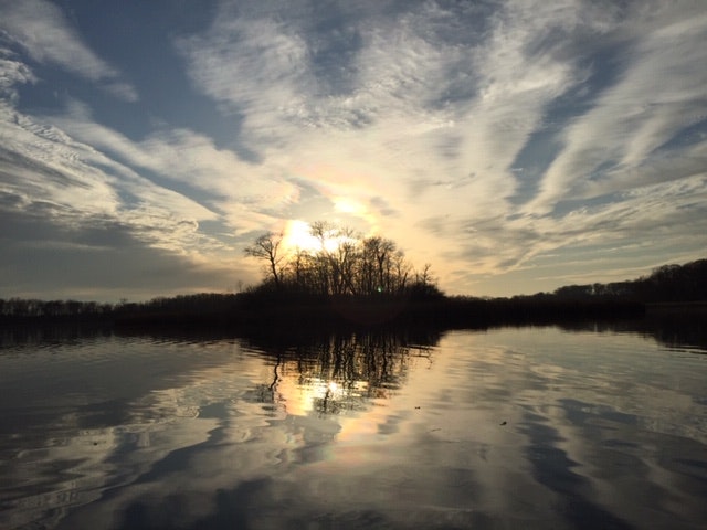 A serene sunset over a calm lake with a reflection of the sky and silhouetted trees.