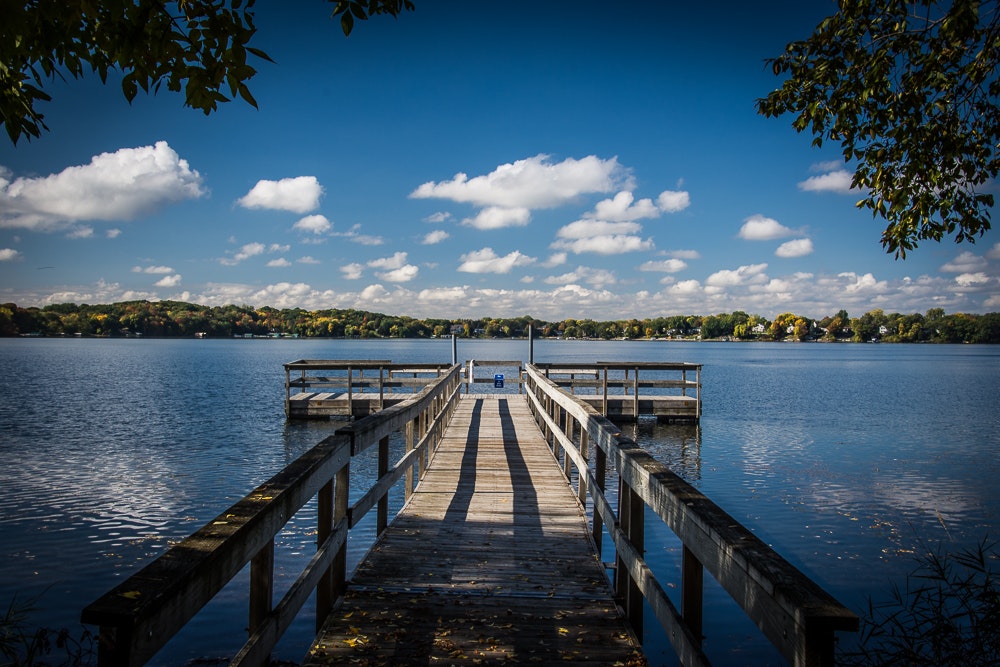 A wooden pier leading to a serene lake with clouds above and trees framing the view.