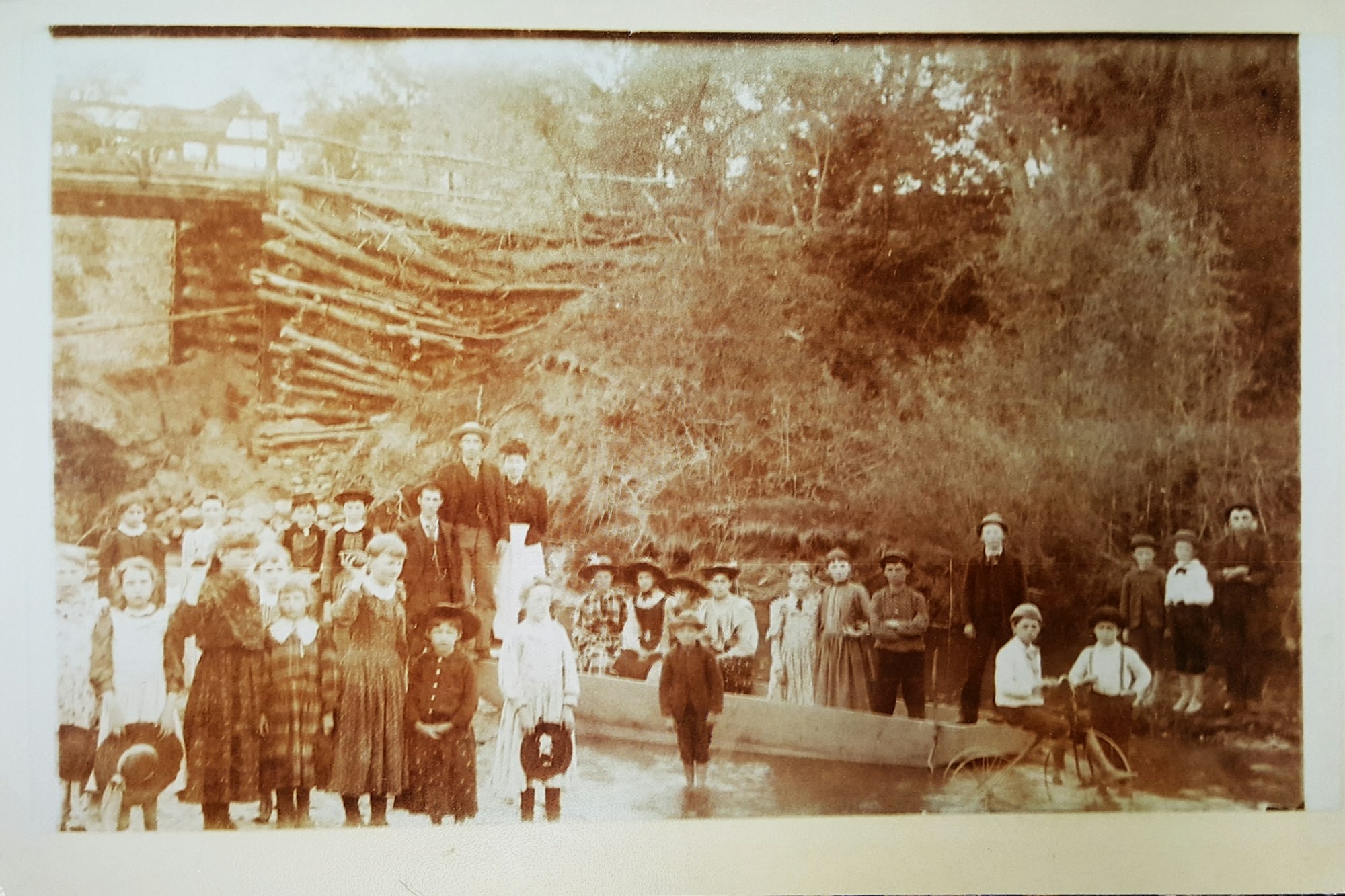 A vintage photo of a group of people outdoors, with children in front and adults behind, near a wooded area and a railing.