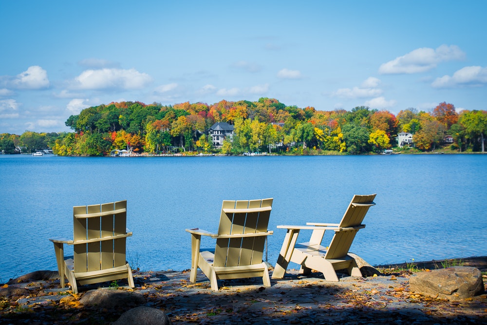 Three Adirondack chairs by a lake with autumn trees in the background.