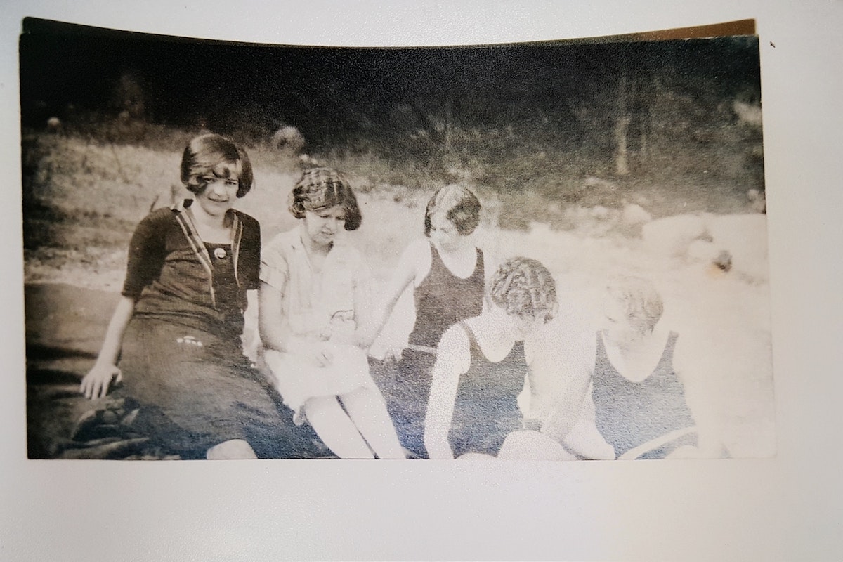 Old photo of five people sitting by water, three looking at camera, two turned away, vintage clothing style.