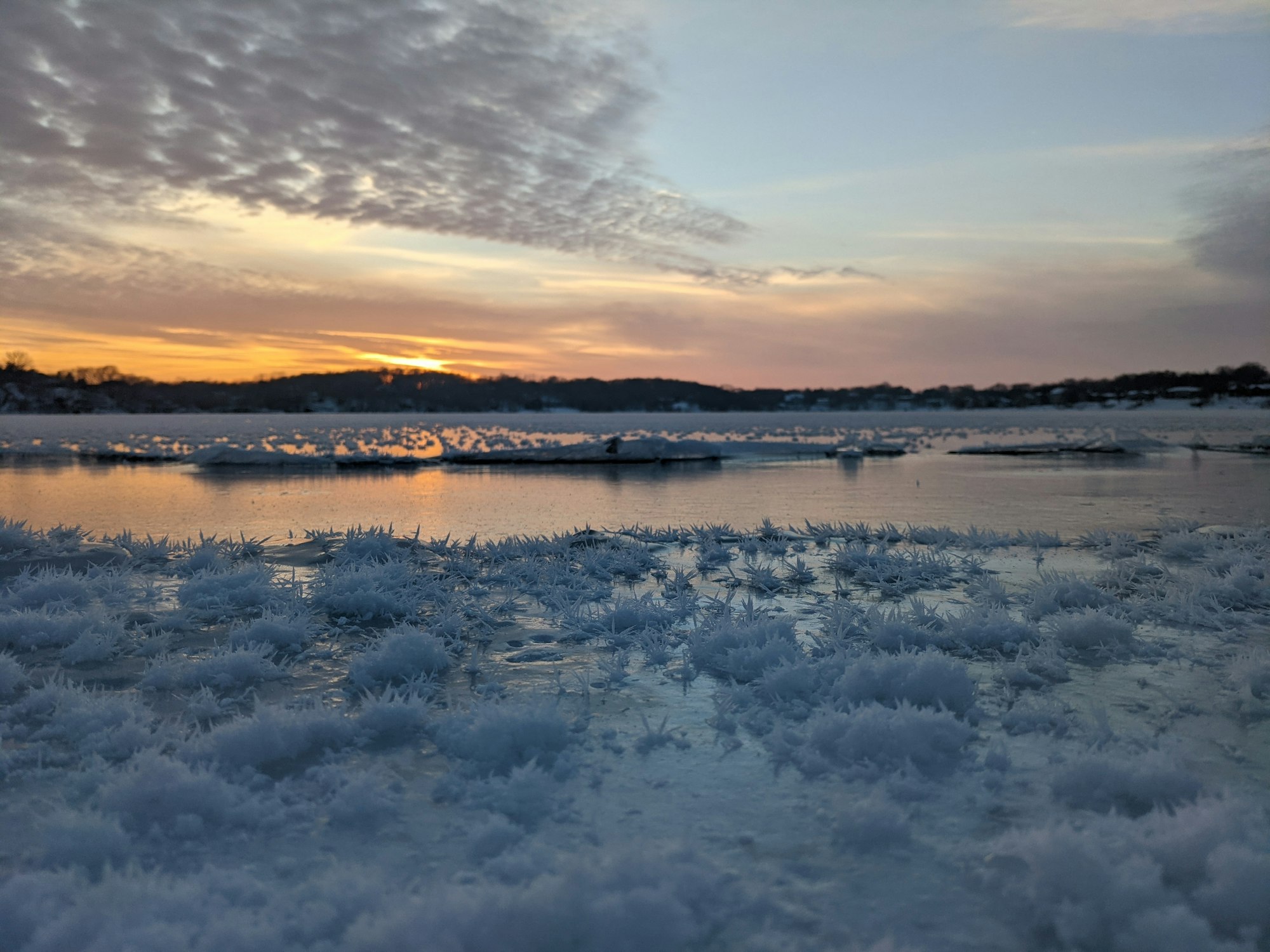 Frosty ice crystals in the foreground with a sunset and partially frozen river in the background.