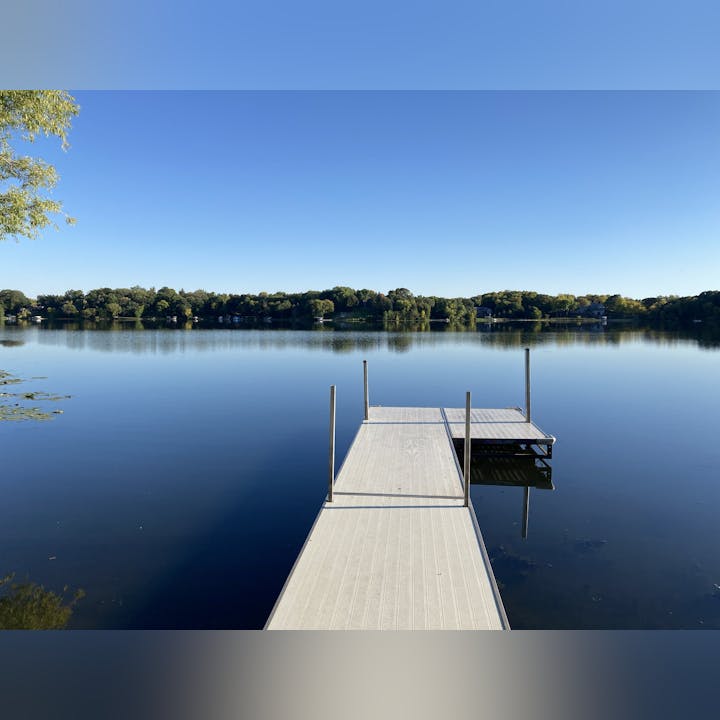 A tranquil lake with a dock extending into the water, surrounded by trees under a clear blue sky.