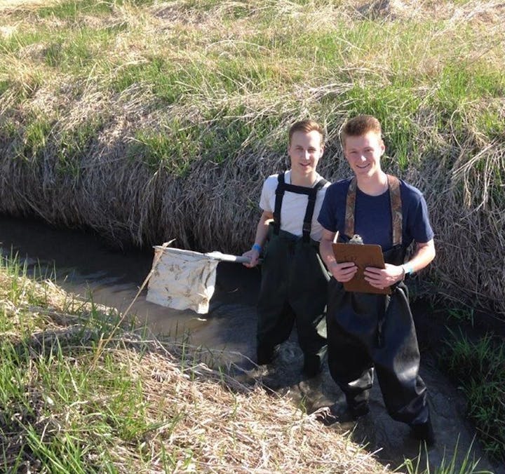 Two individuals in waders with a net and clipboard, possibly conducting fieldwork in a stream.